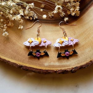May include: A pair of dangle earrings featuring a black bat with a pink rose, hanging from a layered cloud charm with gold accents. The clouds are white and pink.