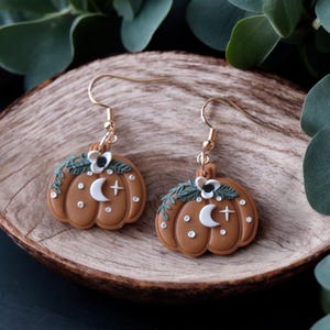 May include: A pair of gold dangle earrings featuring a brown pumpkin with a white crescent moon, a white cross, and white flowers with black centers. The pumpkin is decorated with white dots and green leaves.