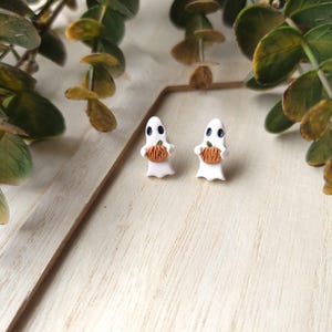 May include: A pair of white ghost-shaped earrings with orange pumpkins in their hands. The earrings are on a white wooden surface.
