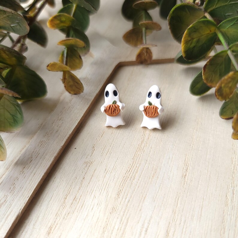 May include: A pair of white ghost-shaped earrings with orange pumpkins in their hands. The earrings are on a white wooden surface.