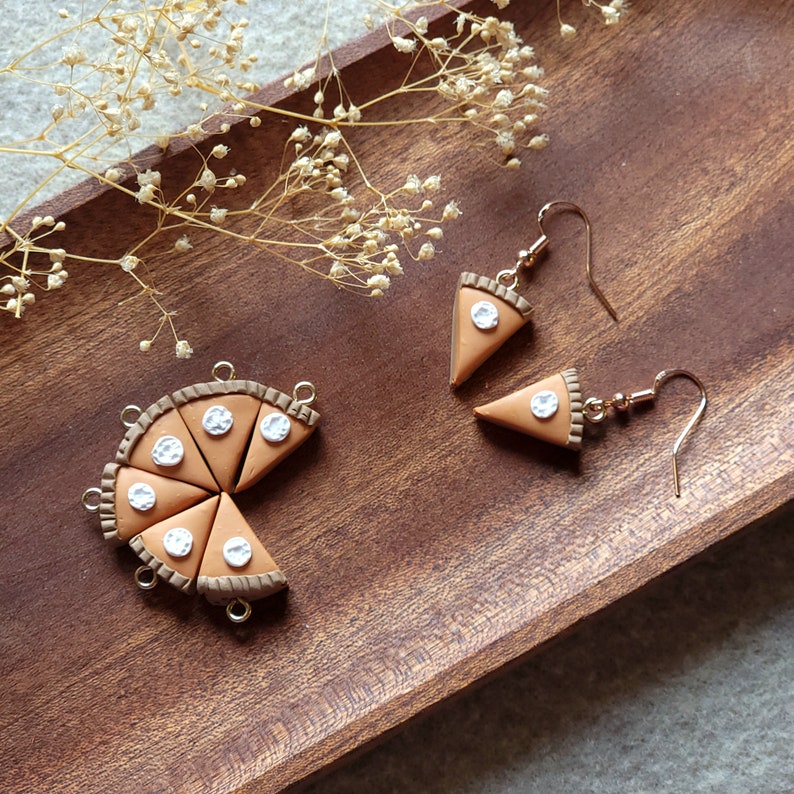 May include: A pair of dangle earrings featuring miniature pie slices with white frosting. The earrings are made of polymer clay and have gold-toned hooks.