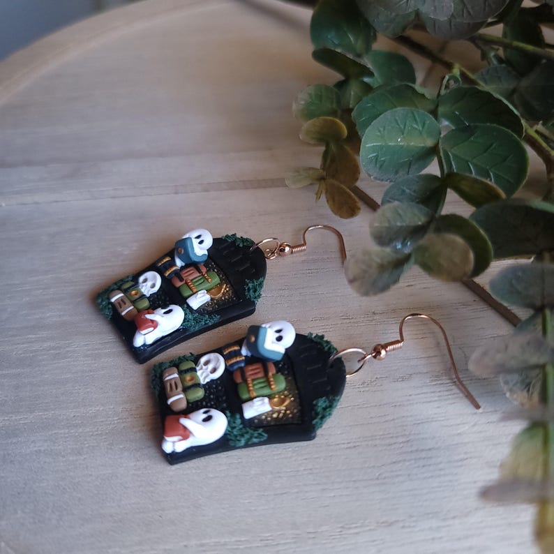 May include: A pair of black and green Halloween-themed earrings featuring a coffin design with ghosts and books. The earrings have gold accents and are made of polymer clay.