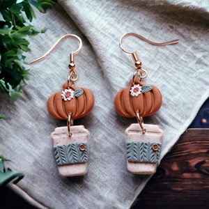 Op de afbeelding: Een paar hangende oorbellen met een bruine pompoen met een witte bloem en groene bladeren, en een witte koffiebeker met een groene en witte gebreide mouw. De oorbellen zijn gemaakt van polymer clay en hebben gouden haakjes.