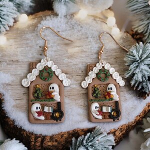 May include: A pair of whimsical gingerbread house earrings. Each earring features a brown house with white icing details, a green wreath, and two friendly ghost figures. One ghost holds a gift, and the other reads a book. Gold-colored ear wires.