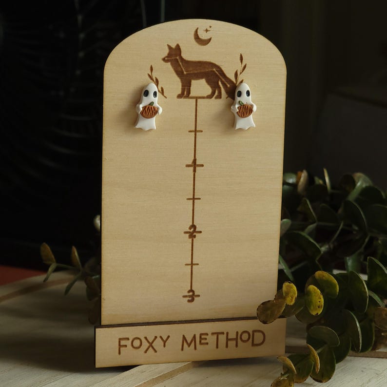 May include: A wooden earring display stand with a fox silhouette and the text "Foxy Method". Two white ghost earrings with orange pumpkins are displayed on the stand.
