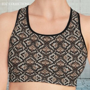 Può includere: Reggiseno sportivo con motivo a pelle di serpente nei toni del marrone, beige e nero. Presenta un design a vogatore con finiture nere intorno allo scollo e alle ascelle. Il logo EDZ COLLECTIONS è visibile in alto a sinistra.