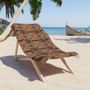 Könnte beinhalten: Ein Holz-Strandstuhl mit einem Stoff mit braunem und schwarzem Schlangenmuster. Der Stuhl steht an einem Sandstrand, im Hintergrund ein Boot und Palmen. Das Meer ist in der Ferne zu sehen.