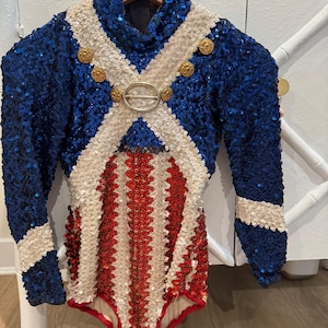 May include: A long-sleeved leotard covered in blue, white, and red sequins. The garment features a high neck, a white sequined cross-body strap design, and gold-toned buttons. The lower section displays vertical red and white sequined stripes.
