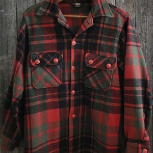 May include: A long-sleeve button-up shirt featuring a plaid pattern in shades of red, black, and olive green. The shirt includes a pointed collar, two chest pockets with buttoned flaps, and matching red buttons down the front placket and on the cuffs.