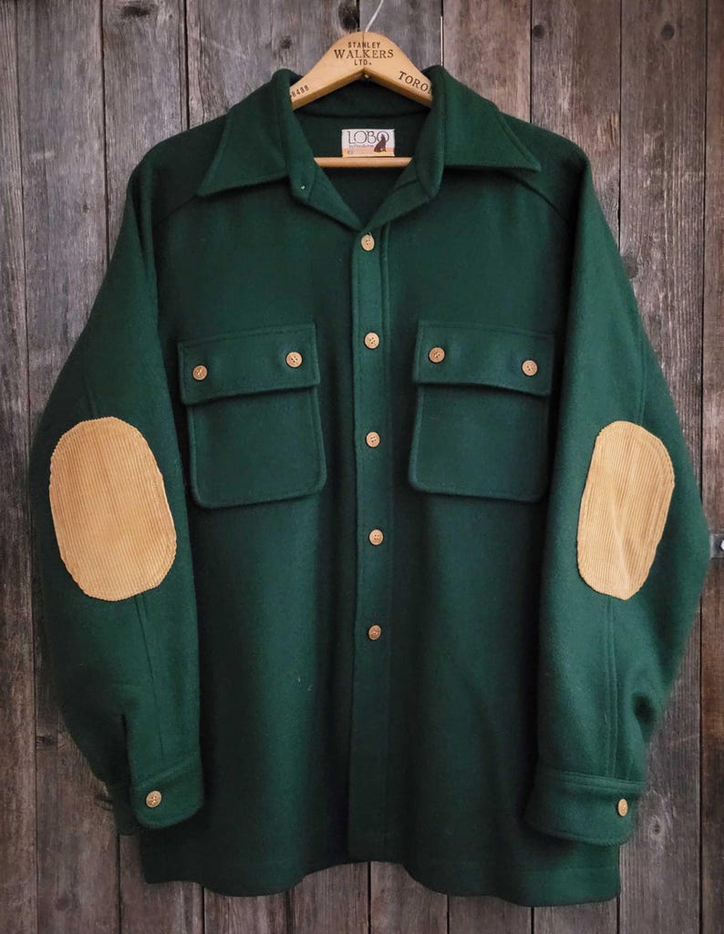 May include: A dark green long-sleeve shirt featuring a pointed collar, a front button closure, and two chest pockets with buttoned flaps. The sleeves include tan corduroy elbow patches, and the interior label displays the text Lobo.