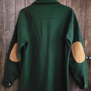 May include: A dark green long-sleeve jacket featuring a collar, buttoned cuffs, and tan corduroy elbow patches. The garment is displayed on a wooden hanger, showcasing a back view with a horizontal seam across the upper shoulders.