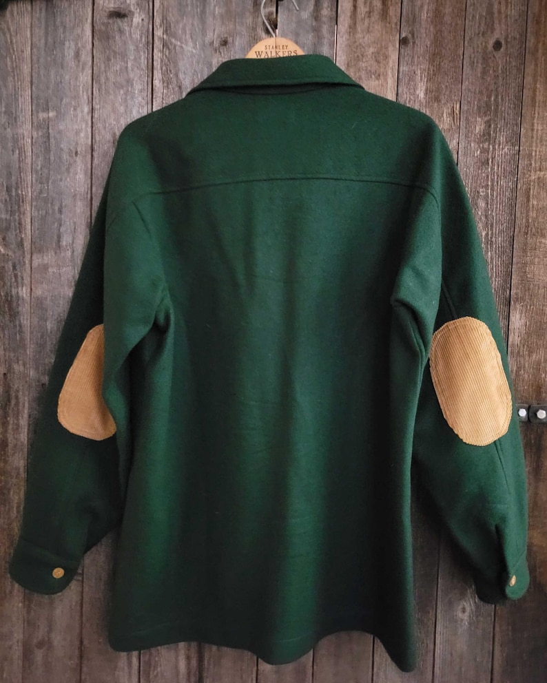 May include: A dark green long-sleeve jacket featuring a collar, buttoned cuffs, and tan corduroy elbow patches. The garment is displayed on a wooden hanger, showcasing a back view with a horizontal seam across the upper shoulders.