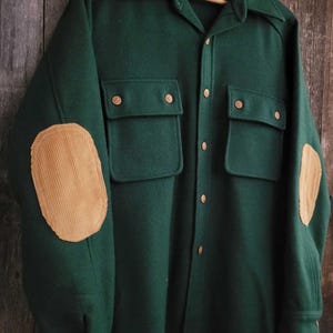 May include: A dark green long-sleeve shirt featuring a collar, a button-down front, and two chest pockets with button closures. The sleeves are accented with tan corduroy elbow patches. The interior label displays the text Lobo.
