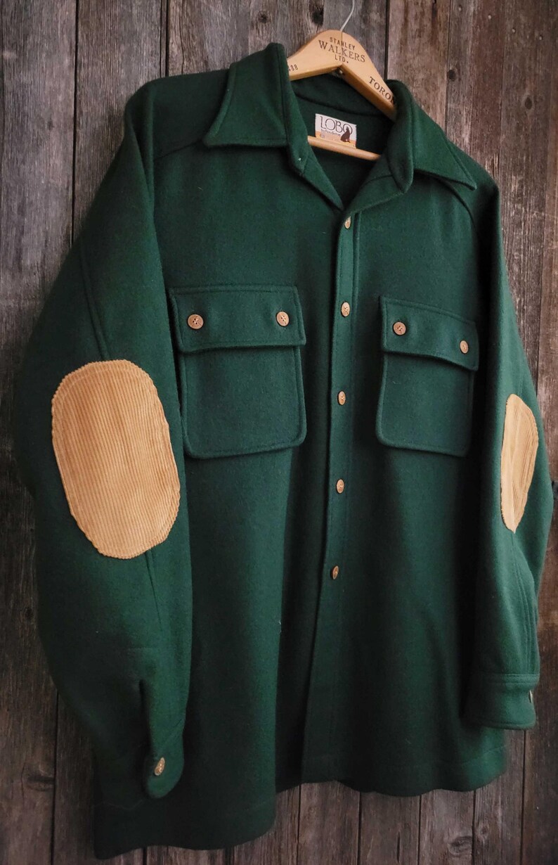 May include: A dark green long-sleeve shirt featuring a collar, a button-down front, and two chest pockets with button closures. The sleeves are accented with tan corduroy elbow patches. The interior label displays the text Lobo.