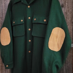 May include: A dark green long-sleeve shirt featuring a collar, a button-down front with light brown buttons, two chest pockets with button flaps, and tan corduroy elbow patches on each sleeve. The interior label displays the text Lobo.