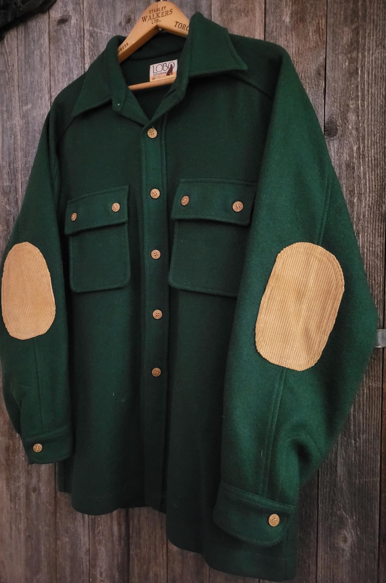 May include: A dark green long-sleeve shirt featuring a collar, a button-down front with light brown buttons, two chest pockets with button flaps, and tan corduroy elbow patches on each sleeve. The interior label displays the text Lobo.