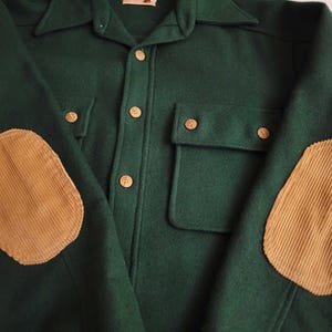 May include: A dark green long-sleeve shirt featuring a button-down front with light brown buttons, two chest pockets with button flaps, and tan corduroy elbow patches on each sleeve. The shirt has a pointed collar and a structured fabric texture.