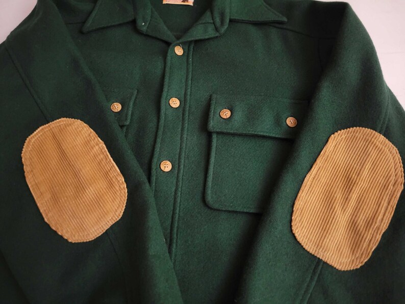 May include: A dark green long-sleeve shirt featuring a button-down front with light brown buttons, two chest pockets with button flaps, and tan corduroy elbow patches on each sleeve. The shirt has a pointed collar and a structured fabric texture.