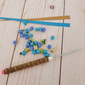 Könnte beinhalten: Eine Sammlung von Bastelmaterialien, darunter blaue und grüne Papierperlen, Papierstreifen und eine Ahle mit Holzgriff und rosa Spitze. Die Perlen sind in verschiedenen Blau- und Grüntönen gehalten. Die Ahle wird zum Basteln und für Perlenarbeiten verwendet.