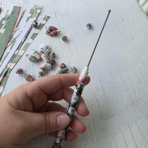 May include: A hand holds a floral-patterned quilling tool with a blue tip, alongside paper strips and rolled paper beads. The tool features a silver metal tip and a handle adorned with pink, white, and blue floral designs.