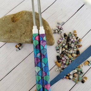 Könnte beinhalten: Zwei silberne Papierperlen-Rollwerkzeuge mit blauer, grüner und violetter Aquarell-Meerjungfrauen-Schuppen-Designs. Die Werkzeuge befinden sich auf einem hellbraunen Stein mit weißem Holz Hintergrund. Es gibt mehrere Papierperlen, die um die Werkzeuge herum verstreut sind.