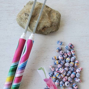 May include: Two bead-making tools with colorful, swirled handles and silver needles. A pile of small, patterned beads in shades of blue, pink, and yellow are scattered nearby, along with a small rock and a piece of paper with a floral design.