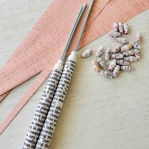 May include: Two white dowel rods wrapped in paper with black text. The text includes the words "they have", "you think", "of what", "to so", "to 80", "of the surface", "they loop", "boldfast", "application", "reach", "been "or", "re defining", "into the", "fig 1 of", "the figure", "ok that", "the word", "re then", "separated", "after", "rs place", "the", "ambered", "though", "have", "ans", "the", "on", "in his", "teks", "or", "of", "as", "mean", "a lake", "ig is", "shoeaning", "meaning", "me", "e are", "af", "just", "reading", "sing", "the", "from", "the", "can", "see", "look", "ating", "to", "f", "g", "first", "leanings", "es", "ago", "owth", "specific", "you", "w", "This", "spe", "of", "seve", "on", "at", "you", "are", "chan".  A pile of colorful paper beads are scattered on a pink and white surface.