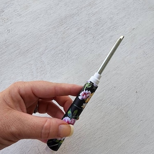 Pode incluir: Um utensílio de metal com padrão floral com fundo branco e design floral preto. O utensílio é segurado na mão.