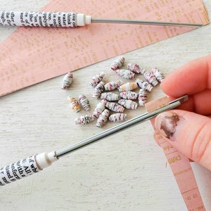 May include: A close-up of a hand threading colourful paper beads onto a metal wire. The beads are made from rolled up pieces of paper with text printed on them. The beads are a mix of pink, white, and orange.