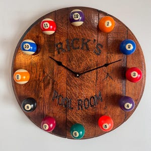 May include: A wooden clock with a pool ball theme. The clock face is made of a dark brown wood with a black clock face and hands. The numbers are represented by pool balls, each with a different color and number. The clock face also has the text "RICK'S POOL ROOM" inscribed on it.