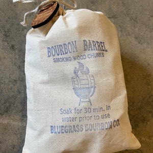 May include: A white canvas bag with a drawstring closure and a wooden tag. The bag is printed with the text "BOURBON BARREL SMOKING WOOD CHUNKS" and a blue illustration of a grill with a barrel on top. The text "Soak for 30 min. in water prior to use BLUEGRASS BOURBON CO." is also printed on the bag.