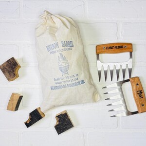 May include: A collection of grilling accessories and wood chunks. Includes a canvas bag of bourbon barrel smoking wood chunks, two meat shredder claws with wooden handles, and several wood chunks. The bag reads "Bourbon Barrel Smoking Wood Chunks".