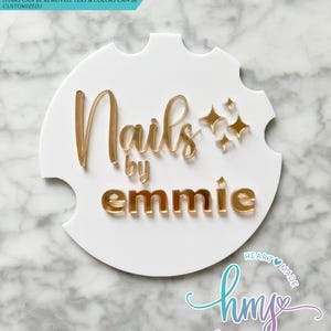 May include: White round sign with gold lettering that reads "Nails by emmie" with gold stars. The sign has a scalloped edge. The text "HEART MADE" and "hmy STUDIOS" is in a purple and blue script font.
