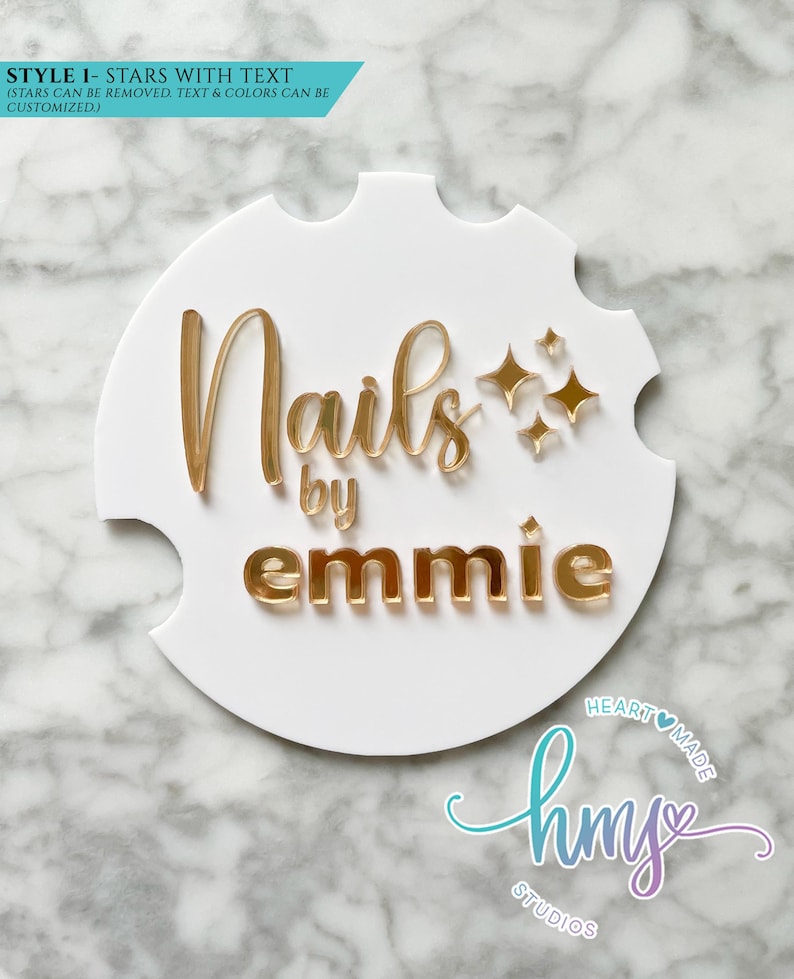 May include: White round sign with gold lettering that reads "Nails by emmie" with gold stars. The sign has a scalloped edge. The text "HEART MADE" and "hmy STUDIOS" is in a purple and blue script font.