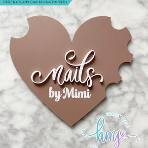 May include: A pink heart-shaped sign with the text "nails by Mimi" in white lettering. The sign also features a smaller logo with the text "hmy studios" in a gradient of blue and purple.