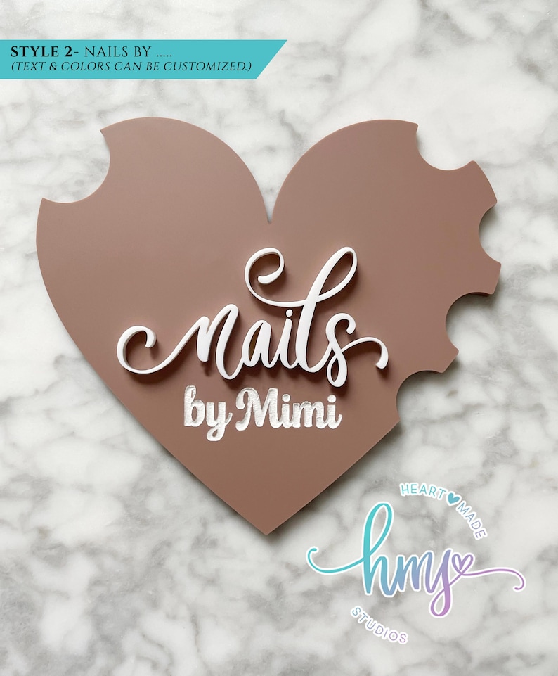 May include: A pink heart-shaped sign with the text "nails by Mimi" in white lettering. The sign also features a smaller logo with the text "hmy studios" in a gradient of blue and purple.