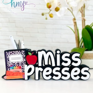 May include: Black desk organizer with a red apple, a notebook, pencils, and crayons. The organizer has the words "Miss Presses" in white letters.