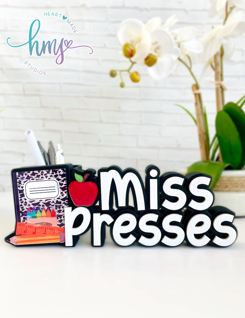 May include: Black desk organizer with a red apple, a notebook, pencils, and crayons. The organizer has the words "Miss Presses" in white letters.