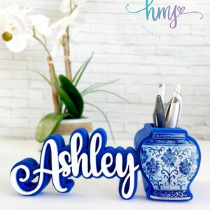 May include: Blue and white personalized desk organizer with the name "Ashley" in white lettering. The organizer features a blue and white porcelain design with floral patterns. The organizer is a great way to keep your desk tidy and organized.