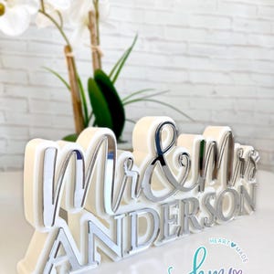 May include: White and silver "Mr. & Mrs. Anderson" wedding sign with a white background. The sign is made of acrylic and has a 3D effect. The sign is decorated with a heart and the text "hmy studios" in a script font.