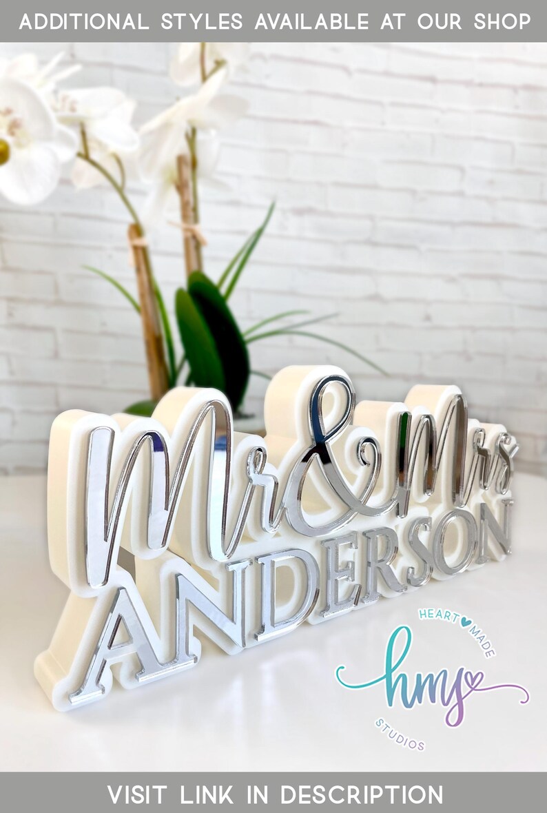 May include: White and silver "Mr. & Mrs. Anderson" wedding sign with a white background. The sign is made of acrylic and has a 3D effect. The sign is decorated with a heart and the text "hmy studios" in a script font.