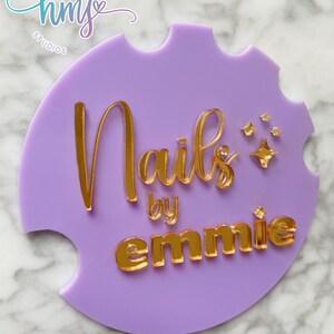 May include: Purple acrylic sign with gold lettering that reads "Nails by emmie" with a small gold star design. The sign has a heart-shaped logo in the top left corner that reads "hmy studios" with a heart.