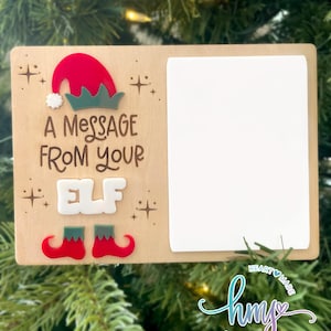 Pode incluir: Enfeite de Natal de madeira com o texto "A Message From Your Elf". O enfeite apresenta um chapéu de elfo vermelho, uma gola verde e sapatos de elfo vermelhos. Possui um espaço retangular branco para uma foto.