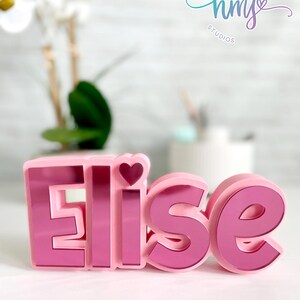 May include: Pink 3D name sign with the name "Elise" spelled out in large letters. The letters are pink with a mirrored finish. A small pink heart is on top of the "E". The sign is on a white surface.  HEART MADE hm STUDIOS VISIT LINK IN DESCRIPTION
