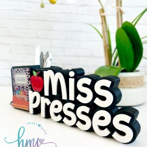 May include: Black and white desk organizer with the words "miss presses" in white letters. The organizer has a red apple and a small stack of books with a leopard print cover. The organizer is on a white surface with a white and green plant in the background.
