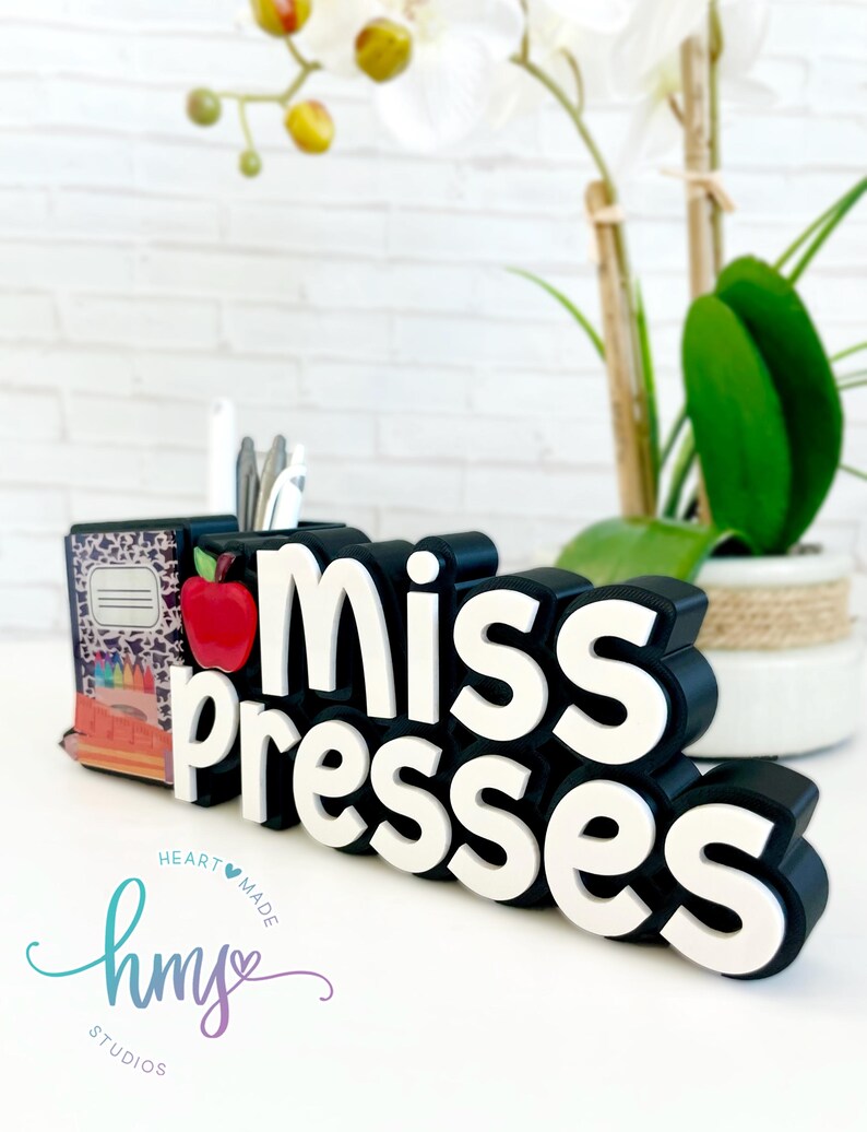 May include: Black and white desk organizer with the words "miss presses" in white letters. The organizer has a red apple and a small stack of books with a leopard print cover. The organizer is on a white surface with a white and green plant in the background.