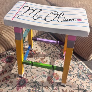 May include: A wooden stool with a seat painted like notebook paper featuring the text Mrs. Oliver and a small pink heart. The four legs are painted to resemble yellow pencils with pink erasers, while the support rungs are colored like crayons.