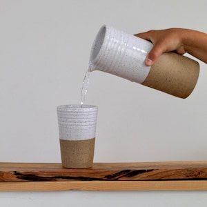 May include: A white and brown speckled ceramic pitcher pouring water into a white and brown speckled ceramic mug. The pitcher and mug are both on a wooden surface.