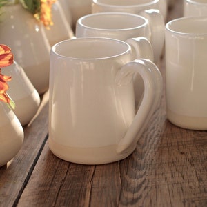 May include: A collection of off-white ceramic mugs with curved handles, arranged on a rustic wooden surface. The mugs have a simple, elegant design, with a slightly darker base. Some vases with flowers are in the background.