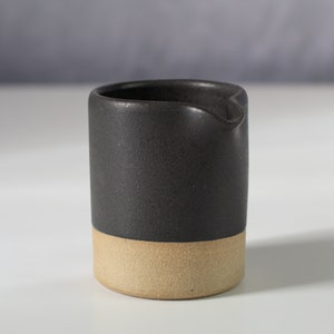 May include: A black ceramic creamer with a light brown base. The creamer has a rounded shape and a small spout.